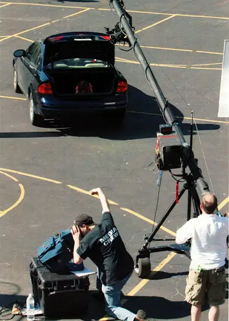 media crew filming a person sitting in the the trunk of a car that is sitting on a blacktop